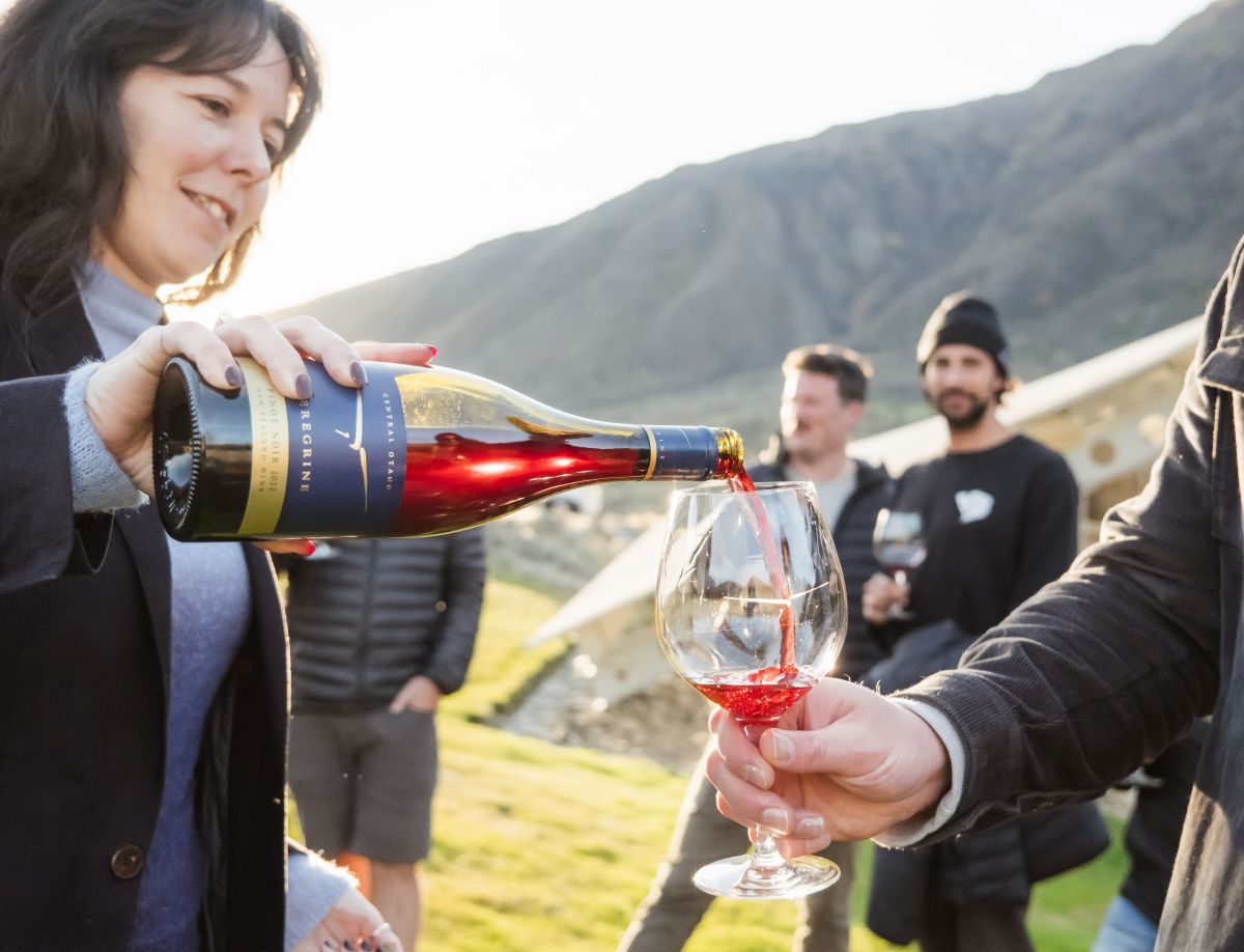 Altitude Tours' wine tour experience at Peregrine Wines. Person pouring a glass of red wine whilst another person holds the glass. Group seen in the background.