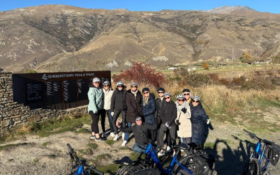group on a Better by Bike tour posing for a photo in front of the Queenstown Trail Trust sign.