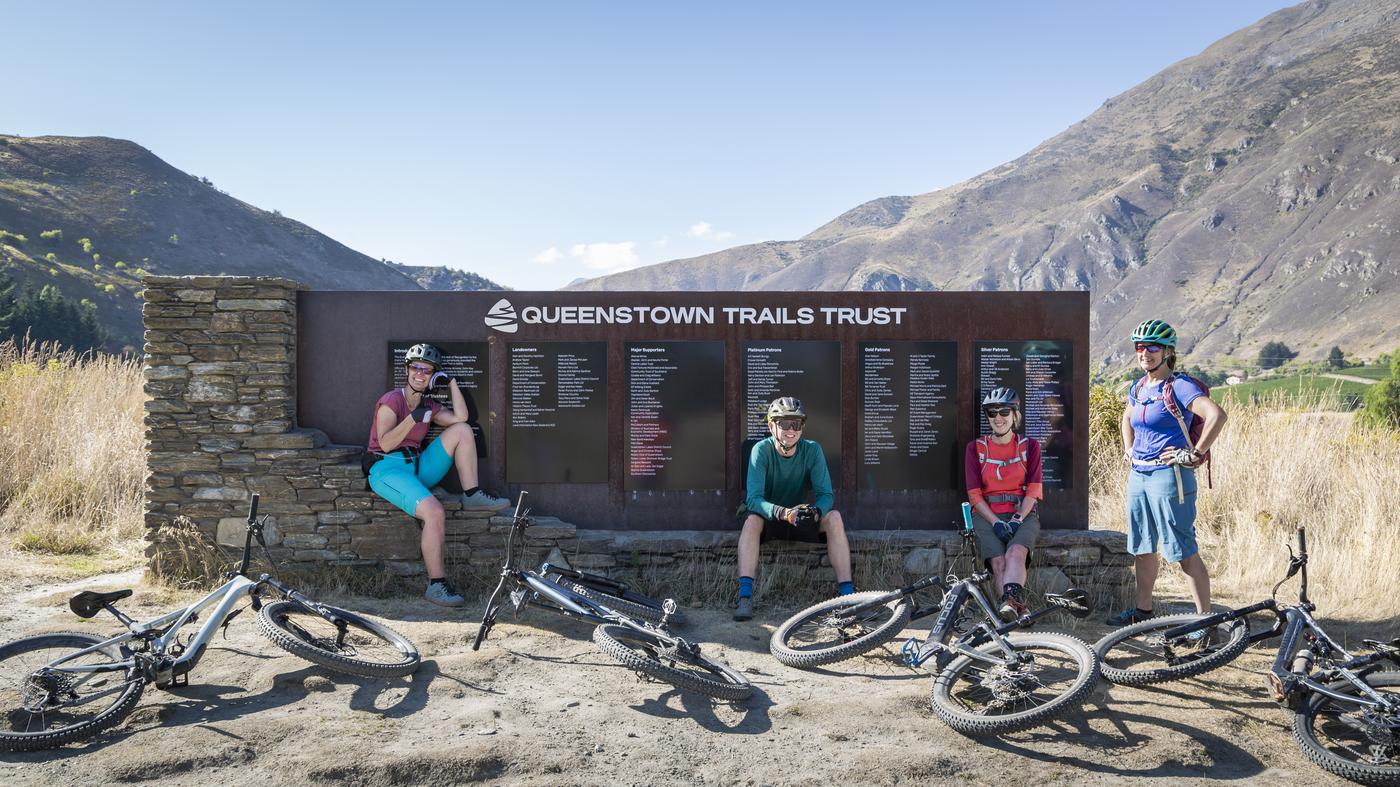 Four people taking a break from riding on the Arrow River Bridges trail in front of the Queenstown Trails Trust sign