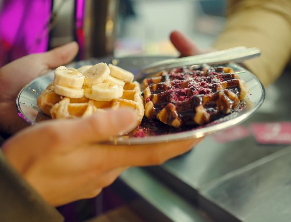 Close up of person giving two waffles on a plate to someone else. One waffle has bananas on top, the other has jam.