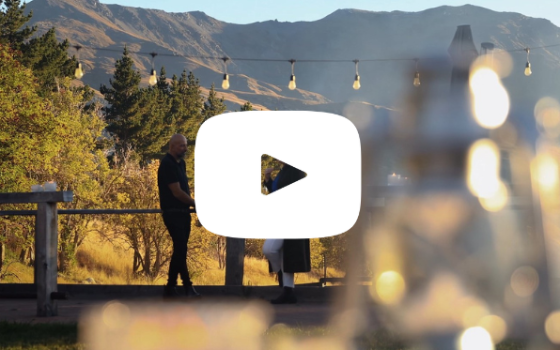 A still from 'Your Ultimate Business Event Destination' YouTube video of two people talking at Royalburn Station with montains in the background. YouTube play symbol in the centre.