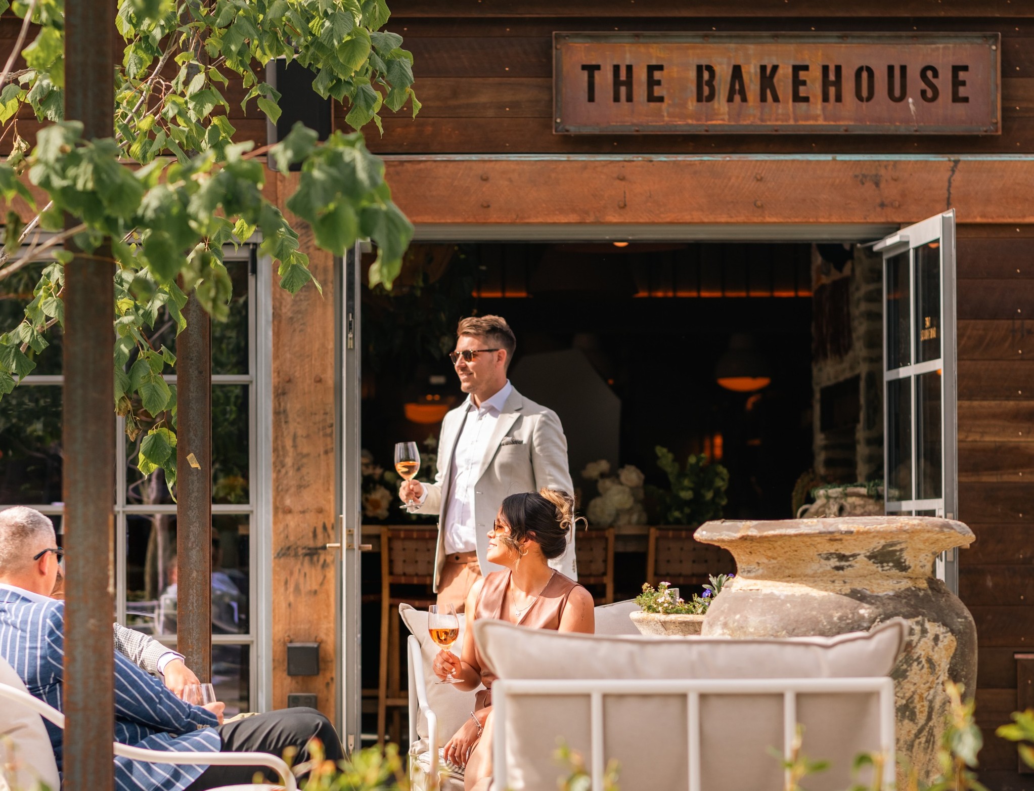 3 people enjoying the outside space of Ayrburn's venue, The Bakehouse. Man holding a glass of wine, the other 2 sitting on the outdoor furniture. 